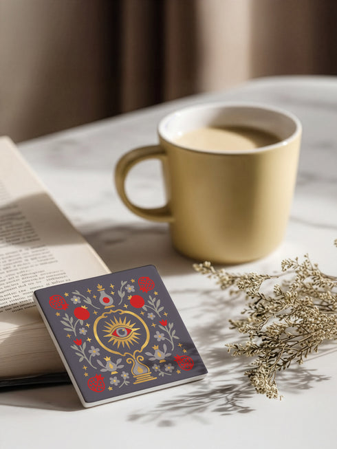 A square ceramic drink coaster with a smooth, matte charcoal grey finish. The surface features a symmetrical, intricate design in metallic gold, deep garnet red, and light grey. In the center is a golden All-Seeing Eye with a red iris, radiating light. The eye is framed by a golden circle and flanked by botanical elements, including red pomegranates and grey flowers with gold details. The design has a folk art, gothic botanical style.
